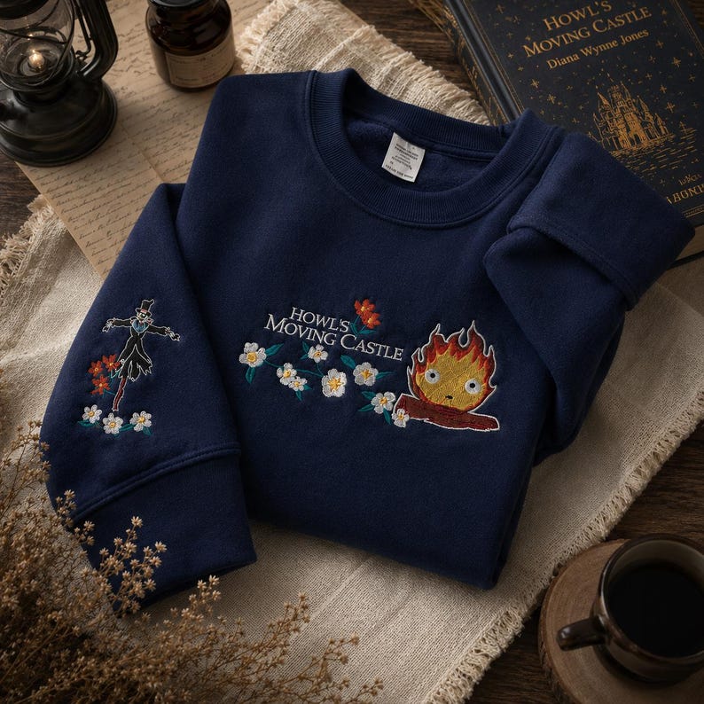 May include: Navy blue sweatshirt with embroidered designs from "Howl's Moving Castle." Features a scarecrow on one sleeve and a fire demon with the text "Howl's Moving Castle" on the chest, surrounded by flowers. The sweatshirt is folded on a neutral-colored cloth.