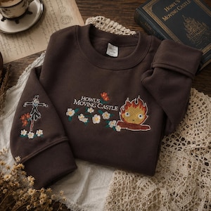 May include: A brown sweatshirt with embroidered designs from "Howl's Moving Castle." The design includes the text "Howl's Moving Castle," a fire demon, and a scarecrow. The sweatshirt is on a white lace doily.
