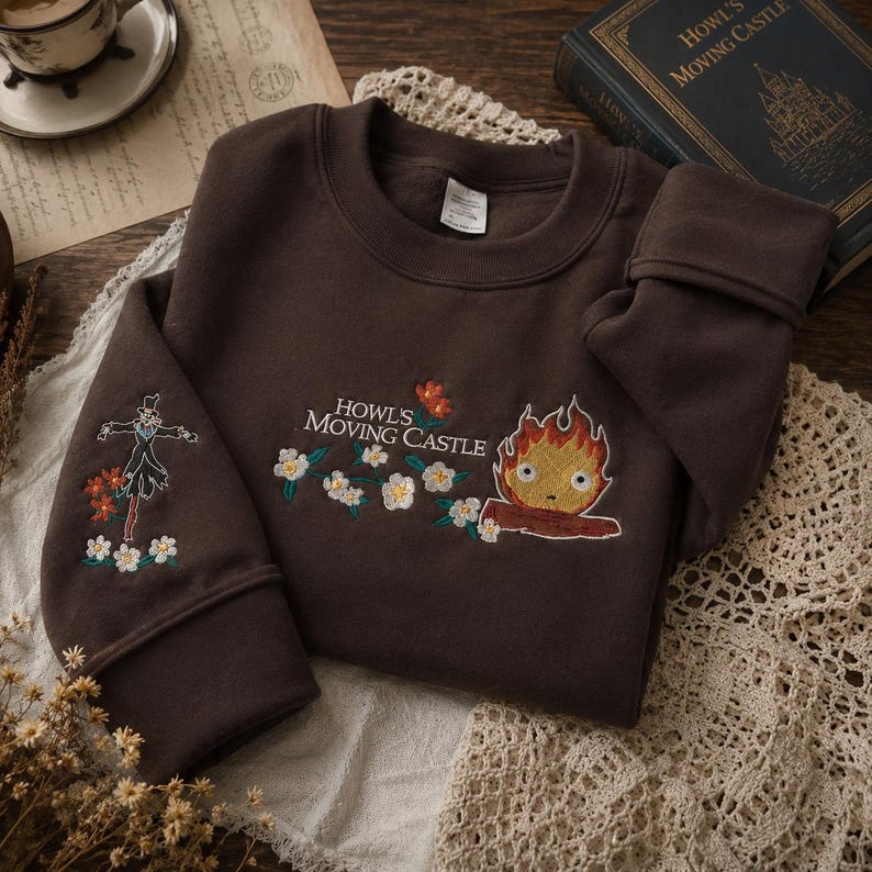 May include: A brown sweatshirt with embroidered designs from "Howl's Moving Castle." The design includes the text "Howl's Moving Castle," a fire demon, and a scarecrow. The sweatshirt is on a white lace doily.