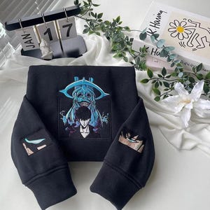 Op de afbeelding: Zwart sweatshirt met geborduurde anime-stijl artwork. Het hoofdontwerp toont een personage met een blauw getint monster en een persoon. De mouwen hebben bijpassende oogontwerpen. Gedetailleerd ontwerp.