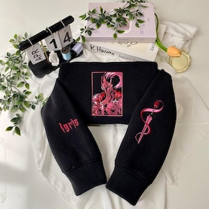 Op de afbeelding: Zwart cropped sweatshirt met rode borduursels. De voorkant heeft een omlijste illustratie van een figuur. De linkermouw heeft het woord "loris" in het rood geborduurd, en de rechtermouw heeft een rood ontwerp.