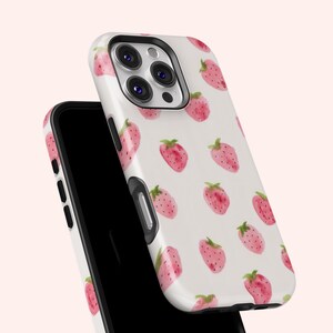 May include: White phone case with a pattern of watercolor strawberries in shades of pink and green. The case is shown on top of a black phone, with the camera and buttons visible.
