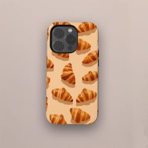 May include: A phone case featuring a pattern of golden-brown croissants on a light orange background. The case has a camera module with three lenses. The design is a repeating pattern of the baked goods.