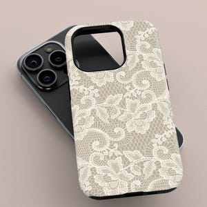 May include: A phone case with a beige background and a white lace pattern. The case is designed to fit a smartphone, with cutouts for the camera and other features. The case is lying on top of a black smartphone.