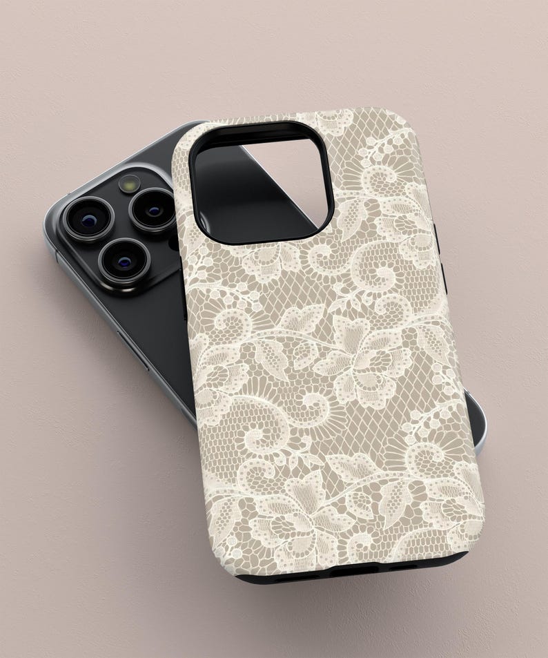 May include: A phone case with a beige background and a white lace pattern. The case is designed to fit a smartphone, with cutouts for the camera and other features. The case is lying on top of a black smartphone.