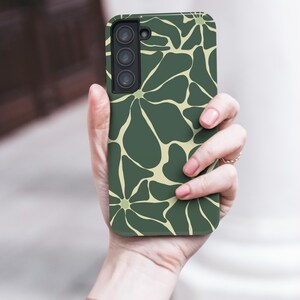 May include: A green phone case featuring a floral design in a lighter green hue. The case is held in a hand, with the phone's camera lenses visible. The case has a matte finish and a contemporary design.