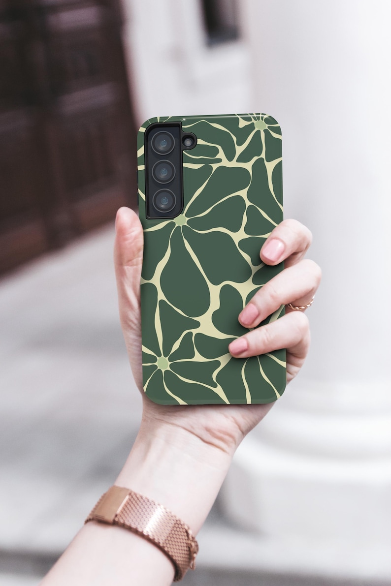 May include: A green phone case featuring a floral design in a lighter green hue. The case is held in a hand, with the phone's camera lenses visible. The case has a matte finish and a contemporary design.