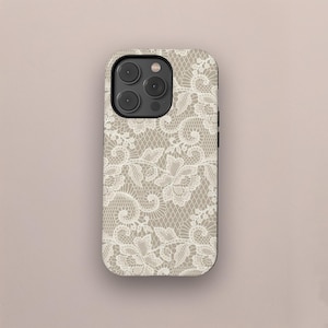 May include: A smartphone case with a white lace design on a tan background. The case has a black border and a camera module with three lenses. The lace pattern features floral and swirling motifs, creating an elegant look.