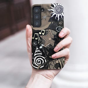 Puede incluir: Una funda de tel&eacute;fono negra y marr&oacute;n con un dise&ntilde;o &uacute;nico que presenta un sol, estrellas, notas musicales y formas abstractas. La funda se sostiene en una mano, mostrando la intrincada obra de arte y la matriz de la c&aacute;mara del tel&eacute;fono.