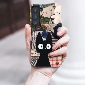 May include: A phone case featuring a black cat illustration with large white eyes. The case has a collage design with a bunny, hearts, and text. The case is held in a person's hand.