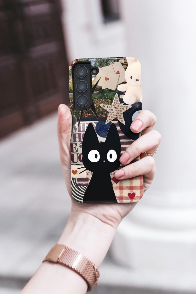 May include: A phone case featuring a black cat illustration with large white eyes. The case has a collage design with a bunny, hearts, and text. The case is held in a person's hand.