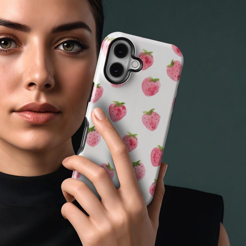 May include: A white phone case featuring a pattern of pink watercolor strawberries with green leaves. The case is held in a person's hand, showcasing the design and the phone's camera.