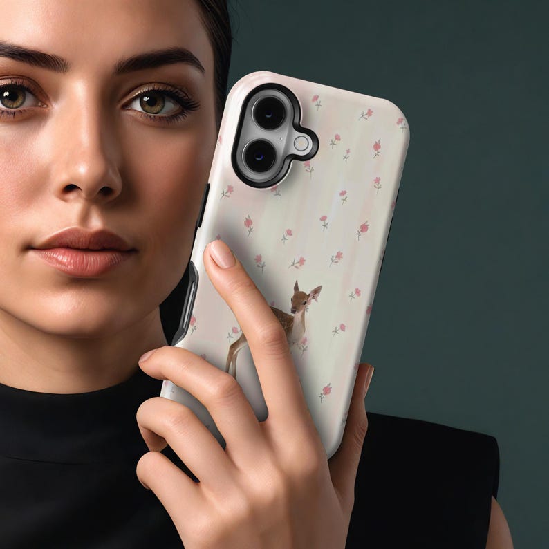 May include: A white phone case with a floral and deer design. The case features a repeating pattern of small pink roses and a watercolor illustration of a deer. The case has a black border and covers the back and sides of the phone.