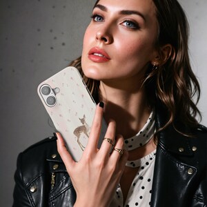 May include: A phone case featuring a watercolour deer illustration and small pink flowers on a light background. The case has a camera cutout. The case is held in front of a person.