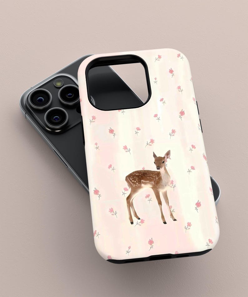 May include: A phone case with a watercolour illustration of a fawn with brown spots and a light pink background. The case is decorated with small pink flowers. The phone case is resting on top of a black phone.