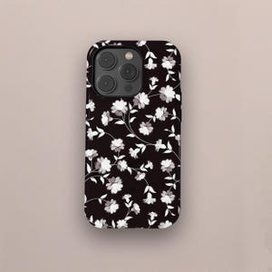 Könnte beinhalten: Schwarze Smartphone-Hülle mit floralem Muster. Das Muster zeigt sich in weißen und grauen Blumen mit grünen Stielen und Blättern. Die Hülle hat eine glänzende Oberfläche und einen Kameraausschnitt.