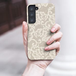 May include: A beige phone case with a delicate, off-white lace pattern. The case has a rectangular shape with a camera module on the upper left. The phone case is held in a hand, showcasing the intricate design.