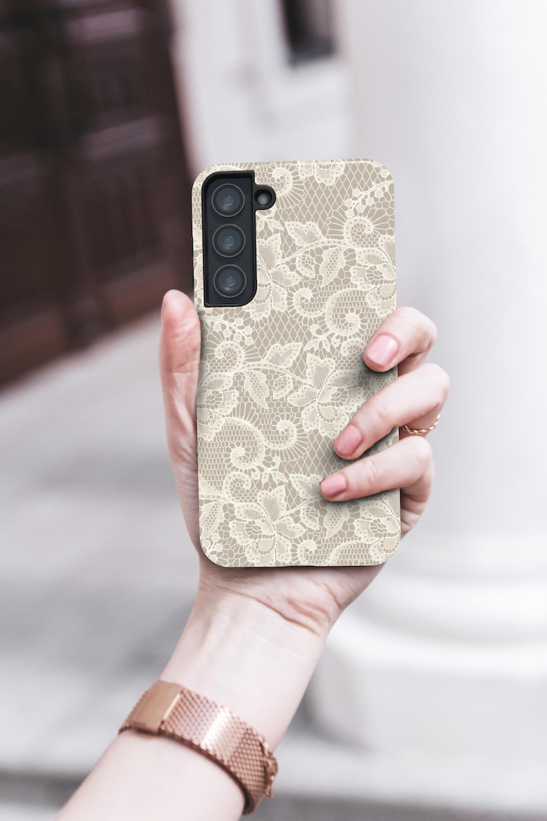 May include: A beige phone case with a delicate, off-white lace pattern. The case has a rectangular shape with a camera module on the upper left. The phone case is held in a hand, showcasing the intricate design.