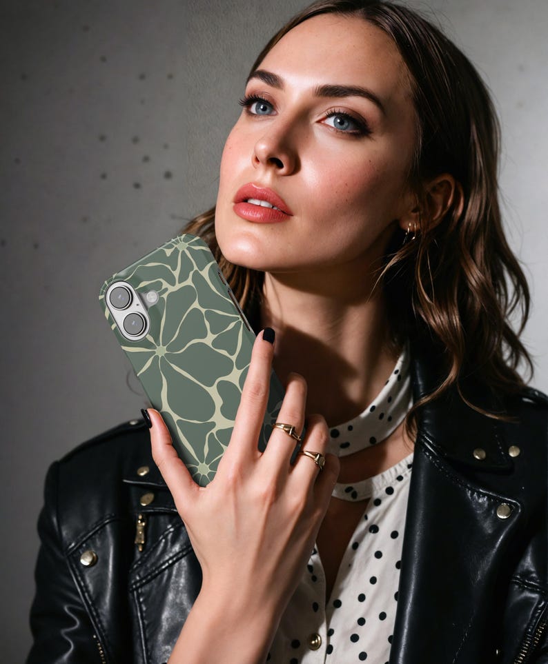 May include: A sage green phone case with a floral design in a lighter beige hue. The case is held by a person wearing a black leather jacket and a white polka dot shirt. The phone case has a dual camera.