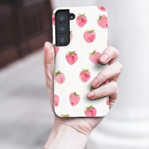 May include: A white phone case featuring a pattern of watercolor strawberries. The strawberries are pink with green leaves and are scattered across the case. The phone case has a camera module with three lenses.