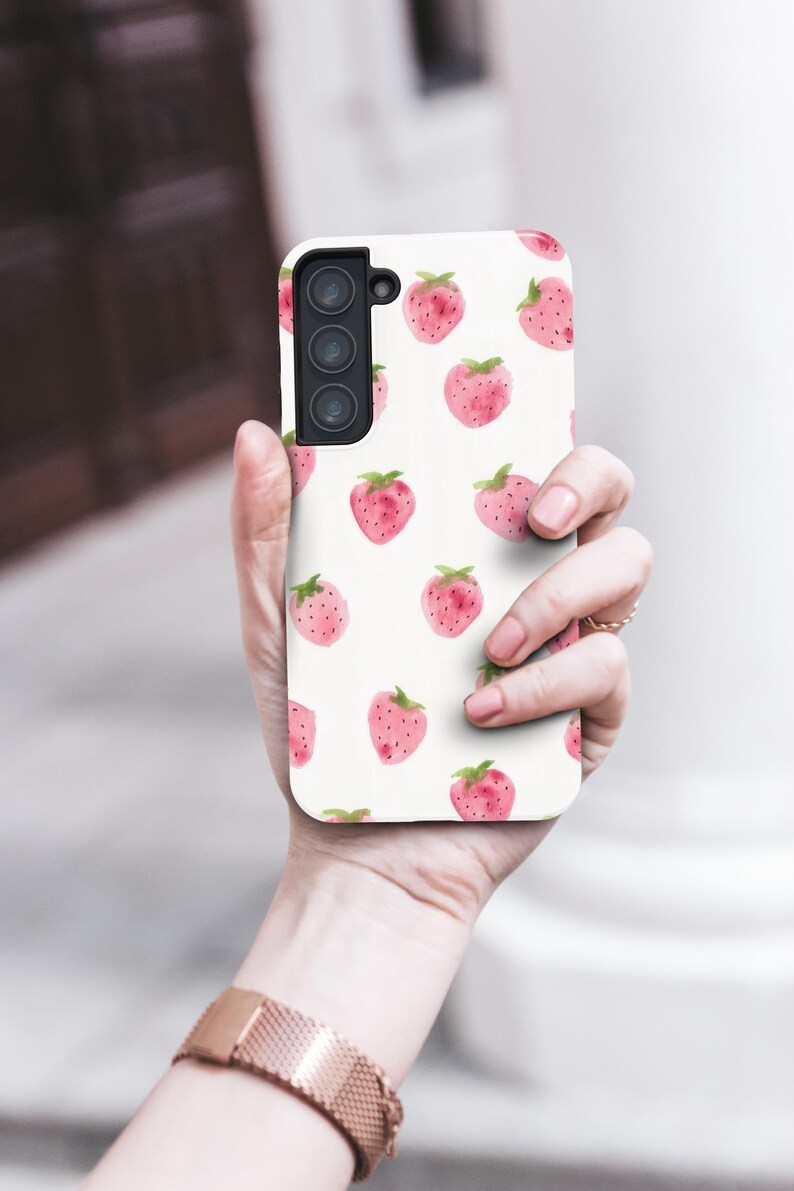May include: A white phone case featuring a pattern of watercolor strawberries. The strawberries are pink with green leaves and are scattered across the case. The phone case has a camera module with three lenses.