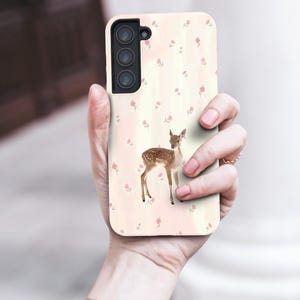 May include: A phone case with a watercolour illustration of a fawn and a floral pattern. The case has a light pink background with small pink flowers. The phone case is held in a person's hand.