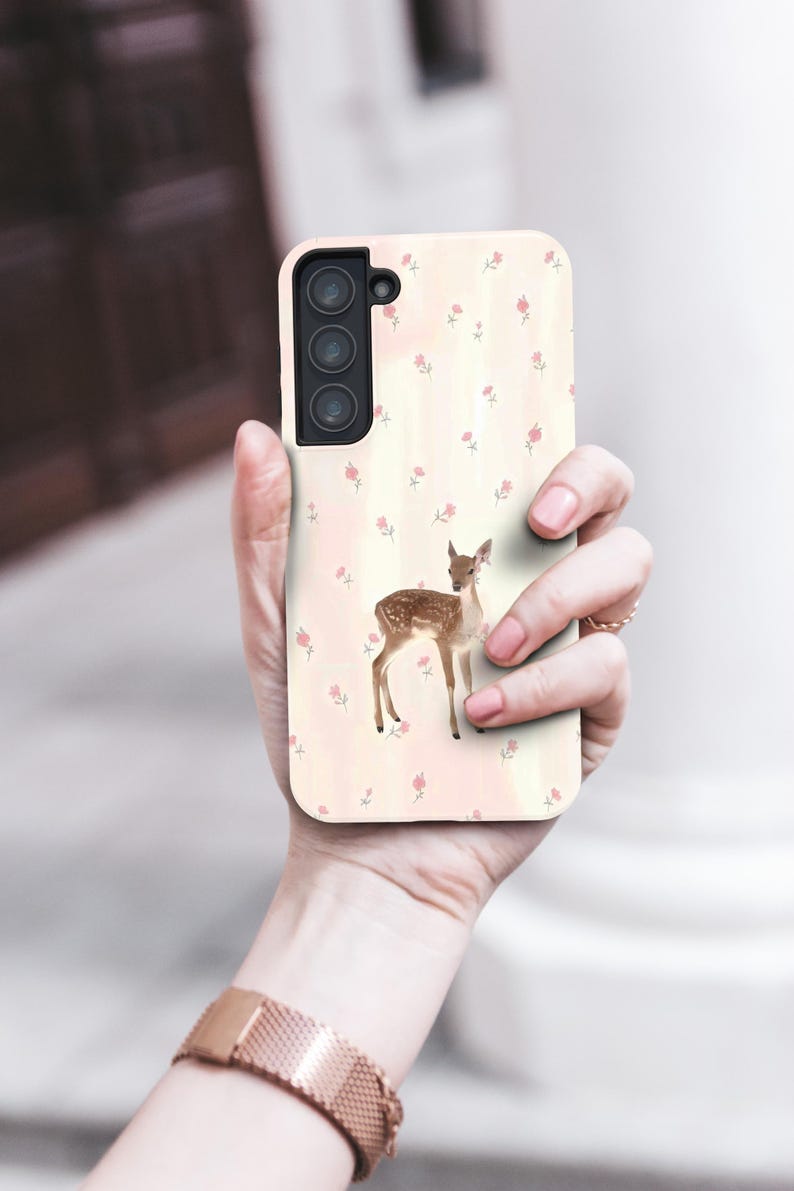 May include: A phone case with a watercolour illustration of a fawn and a floral pattern. The case has a light pink background with small pink flowers. The phone case is held in a person's hand.