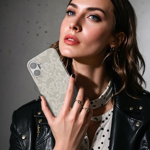 May include: A beige phone case featuring a delicate white lace pattern. The case has a dual-lens camera cutout and a smooth, matte finish. The design is a floral lace overlay, offering a vintage and elegant aesthetic. The case is designed for a smartphone.
