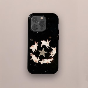 以下が含まれることがあります： 黒い携帯電話ケース。白いウサギと灰色の星が星空の黒い背景に描かれています。ウサギは円形に配置され、キラキラ光るアクセントがあります。ケースは光沢のある仕上がりです。