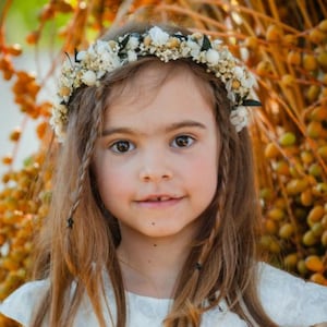 May include: A young girl with long brown hair and a floral crown. She wears a white dress with a floral pattern. The background features a cluster of orange and green berries.