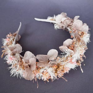 May include: A floral crown featuring dried flowers in shades of beige, cream, and pale pink. The crown is composed of large, delicate petals and smaller, textured blooms, creating a natural and elegant design. The base is a simple white band.
