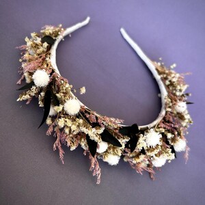 May include: A white headband adorned with dried flowers in shades of cream, pink, and green. The floral arrangement includes small white pom-poms and dark green leaves, with a small black bow. The background is a solid purple.