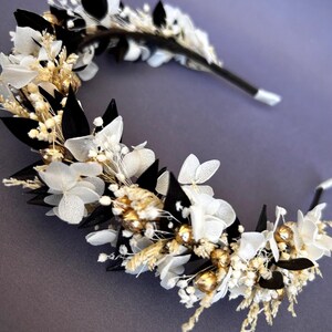 May include: A floral crown and matching hair pin featuring white flowers, black accents, and gold embellishments. The headpiece is set against a gray background, showcasing the delicate details of the handcrafted design.