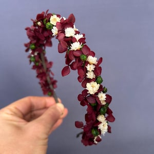 May include: A floral headband featuring burgundy leaves and small white flowers. The headband is adorned with green buds and is held against a muted purple background. This decorative hair accessory is suitable for special occasions.