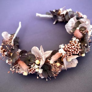 May include: A floral crown featuring a variety of dried flowers in shades of gray, pink, and white. The crown is arranged on a white headband, perfect for weddings or special occasions. The flowers include hydrangeas, small berries, and other delicate blooms.
