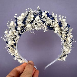 May include: A white and navy blue floral headband made with dried flowers. The headband is made with a white elastic band and is decorated with white and navy blue flowers.
