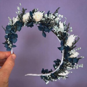 May include: A floral headband featuring white, navy blue, and light green dried flowers and foliage. The headband is held against a purple background. The flowers are arranged in a semi-circle.