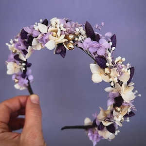 May include: A floral headband featuring a variety of dried flowers in shades of purple, white, and gold. The flowers are arranged on a black headband, creating a delicate and elegant accessory. The headband is held by a hand against a purple background.