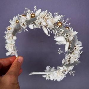 May include: A handcrafted floral crown featuring white dried flowers, gray accents, and gold-colored spherical embellishments. The crown is designed on a white headband, perfect for weddings or special occasions.