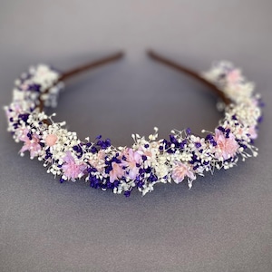 May include: A delicate headband adorned with a circle of dried flowers in shades of white, pink, and purple. The headband is made with a brown wire base.