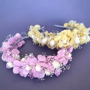 May include: Two floral headbands, one in pale pink and one in cream, with white pom-poms and delicate white sprigs. The headbands are made with dried flowers and are perfect for a wedding or special occasion.