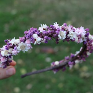 May include: A purple and white flower crown made with dried flowers. The crown is made on a wire base and is decorated with small white flowers and purple flower buds.