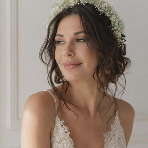 May include: A floral crown composed of small white baby's breath flowers worn atop dark wavy hair. The accessory features a dense arrangement of delicate white blossoms that encircle the head, creating a natural and textured botanical headpiece.