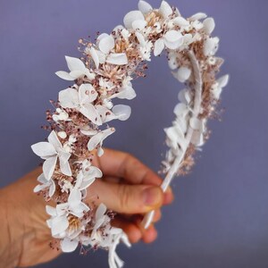 May include: A white floral crown with delicate, layered petals and small, dried flower accents. The crown is held against a purple background, showcasing its intricate design. This handcrafted headpiece is perfect for weddings or special occasions.