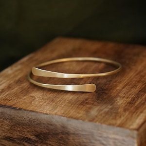 Könnte beinhalten: Ein goldfarbenes Armband aus Metall mit einem Bypass-Design und zwei überlappenden, abgerundeten Enden. Das Armband hat eine glatte, polierte Oberfläche und ein schmales, flexibles Band, das sich zu einer kreisförmigen Form biegt.