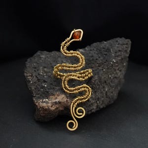 Puede incluir: Un broche de serpiente hecho con alambre dorado que presenta un diseño de cuerpo en espiral y una pequeña piedra de color ámbar engastada en la cabeza. El alambre metálico está intrincadamente enrollado para formar la silueta serpentina.