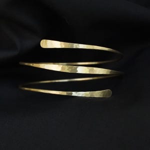 Könnte beinhalten: Ein Oberarmreif aus goldfarbenem Metall mit einem Spiraldesign aus drei Windungen. Die Oberfläche des Metallbandes weist eine gehämmerte Textur auf, die eine Reihe kleiner, reflektierender Vertiefungen über die gesamte Länge erzeugt.