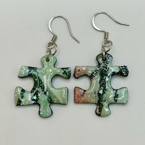 May include: A pair of dangle earrings featuring puzzle piece charms. The charms have a glossy finish with a marbled pattern in shades of sage green, dark green, and hints of peach, accented with fine silver glitter throughout the surface.