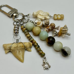May include: A keychain with a silver clasp and various charms. Charms include a shark tooth, small seashells, a carved turtle, and beads in shades of green, brown, and cream. The keychain is on a white background.