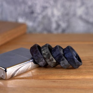 May include: Four black carbon fiber rings stacked on a wooden surface. The rings have a unique, textured pattern and are a dark gray color.